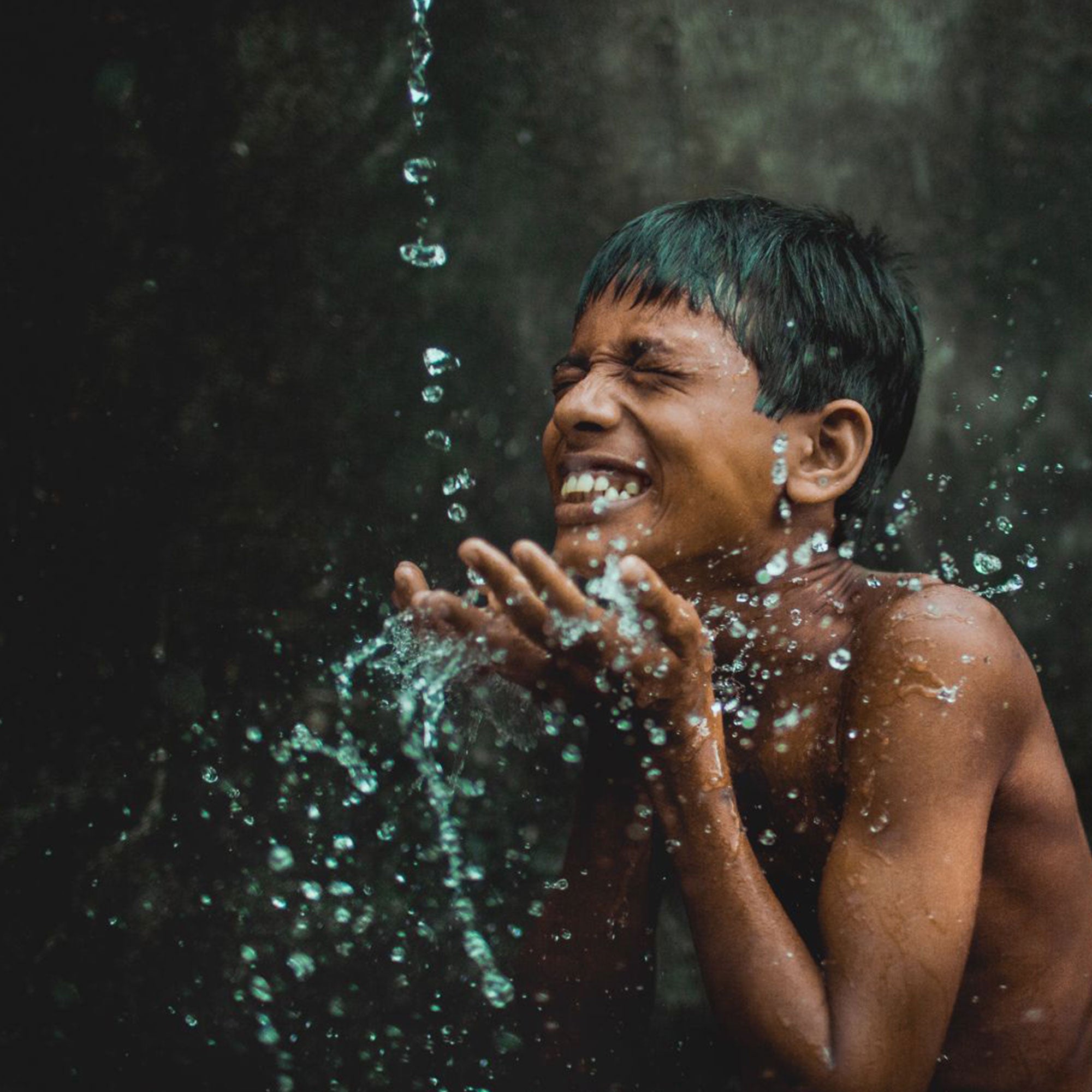 Smiling young child against dark background with water pouring above and splashing onto his face